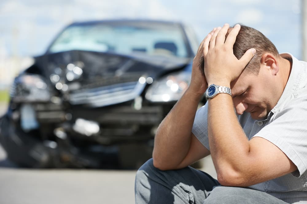 An agitated adult male driver stands in front of his car after a collision in the city road.